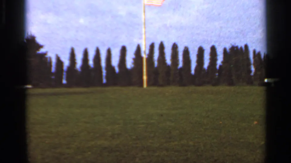 1966 St. Louis-area frame with a flagpole rising from a lawn bordered by dark evergreens and a cloudy sky.