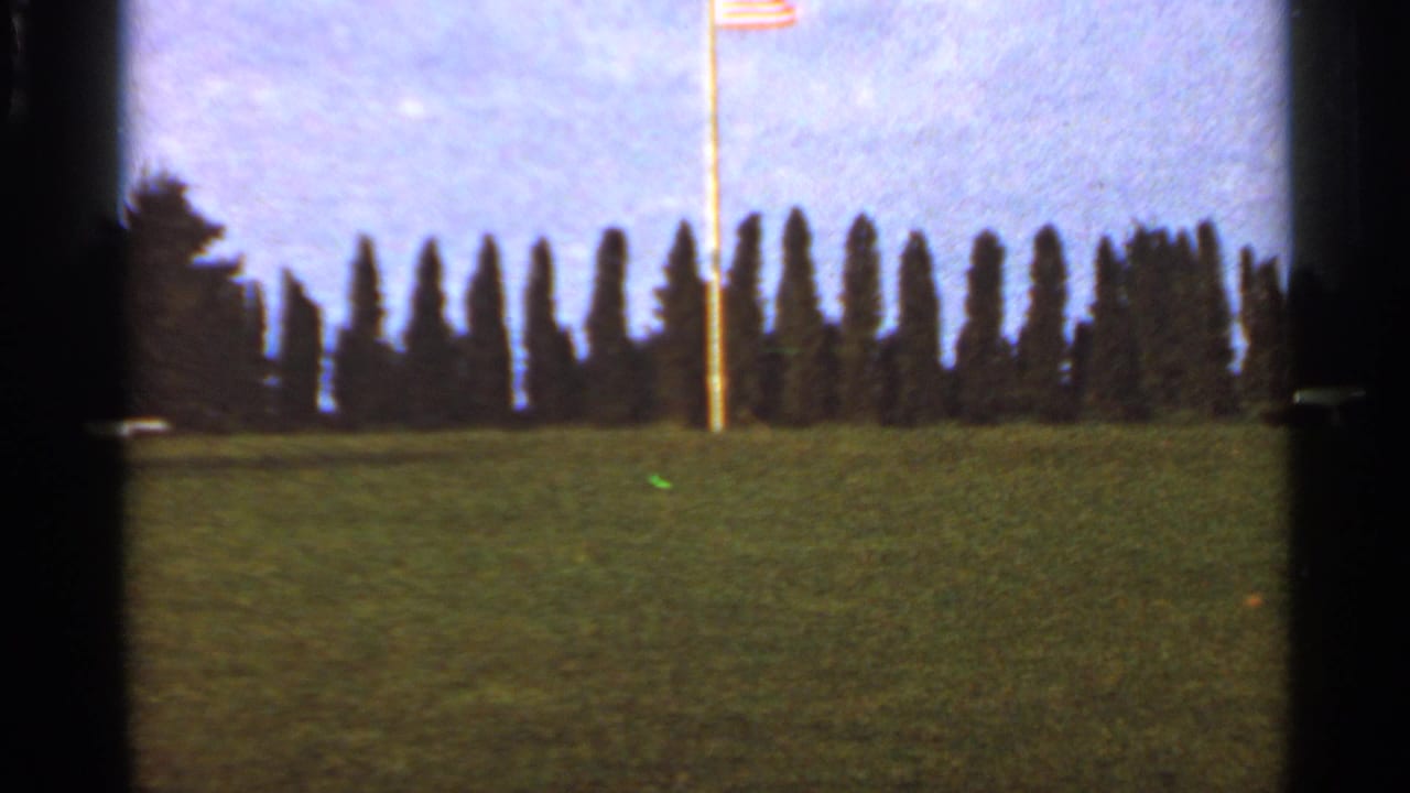 1966 Saint Louis home-movie frame showing an American flag flying above a grassy hill with a dark row of trees behind it.