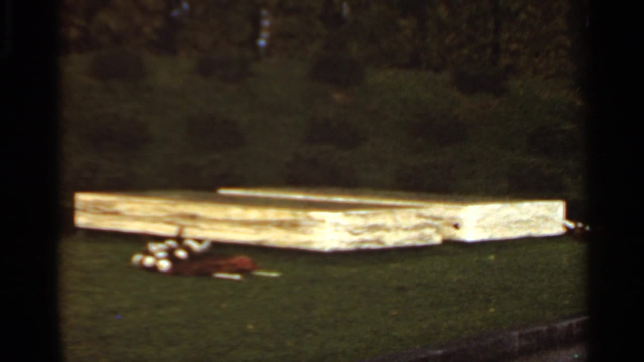 1966 Saint Louis home-movie frame with a low wooden platform on grass, clustered white golf balls, and a brown bag nearby.