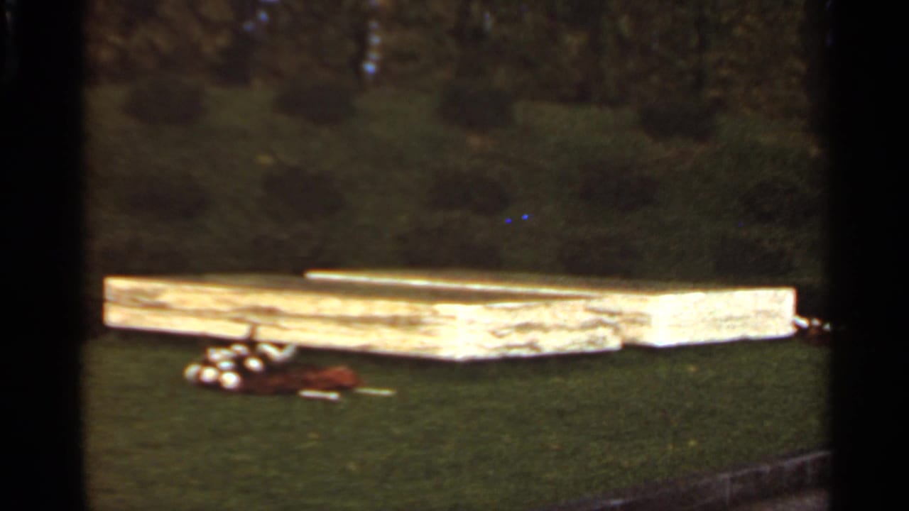 1966 Saint Louis home-movie frame showing a wooden platform across the lawn, with clustered golf balls and a brown bag in the foreground.