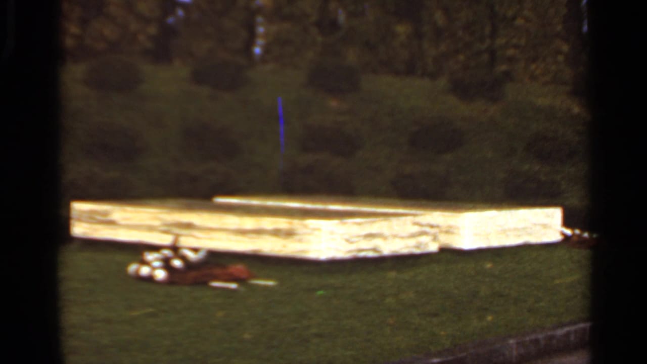 1966 Saint Louis home-movie frame of a wooden platform on a grassy lawn, with golf balls, a brown bag, and dark trees behind.