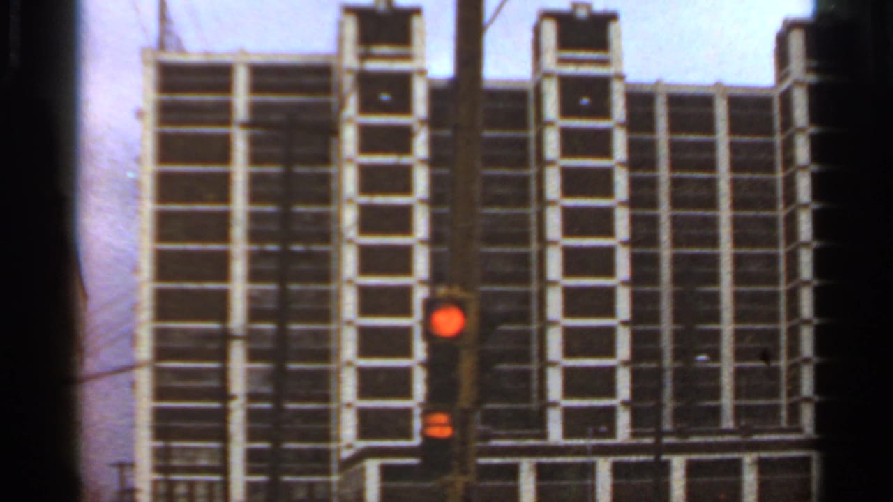 1966 Saint Louis street view with a glowing red traffic light, overhead wires, and a tall white grid-front building in the background.
