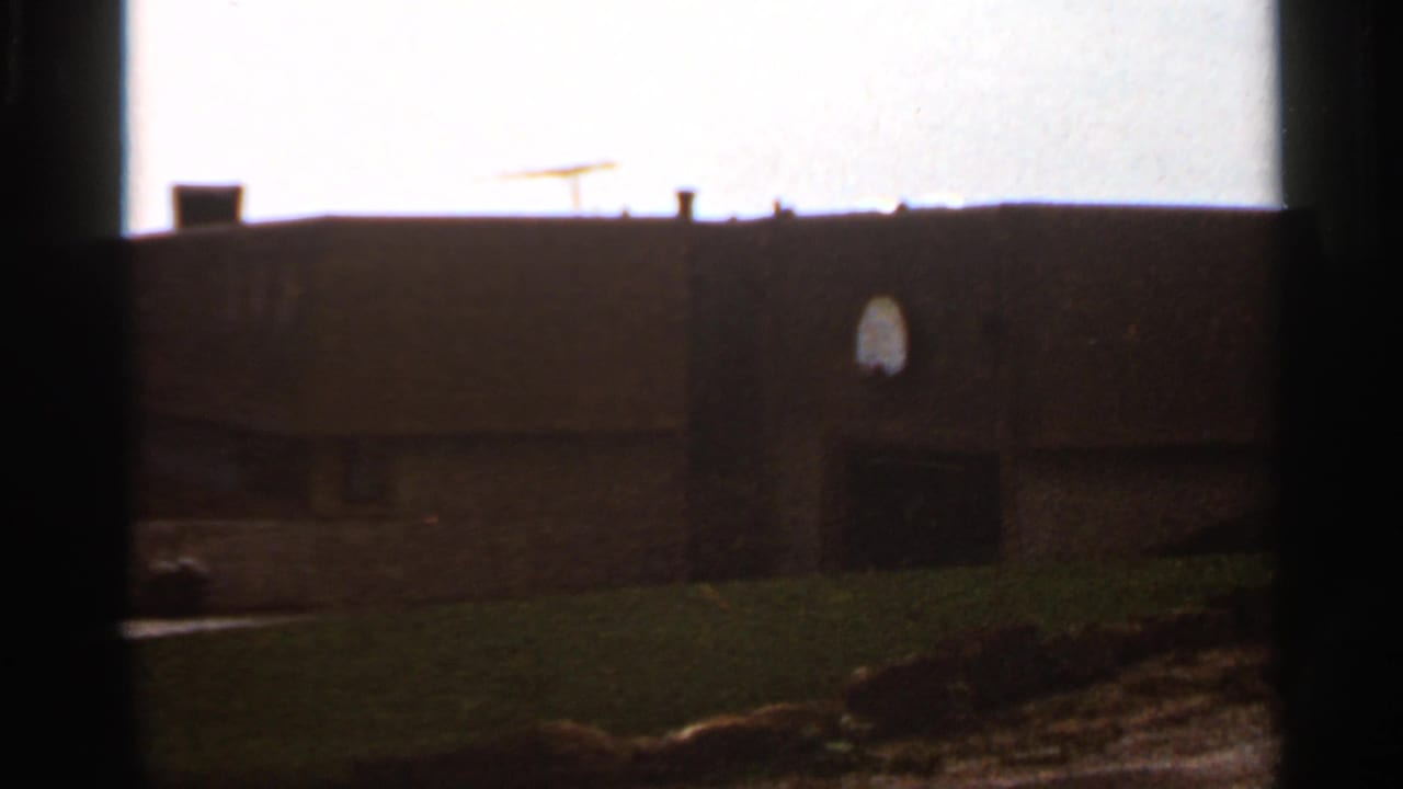 1966 8mm view in Saint Louis showing a dark building facade, roofline, small arched window, and grassy hill under a bright sky.