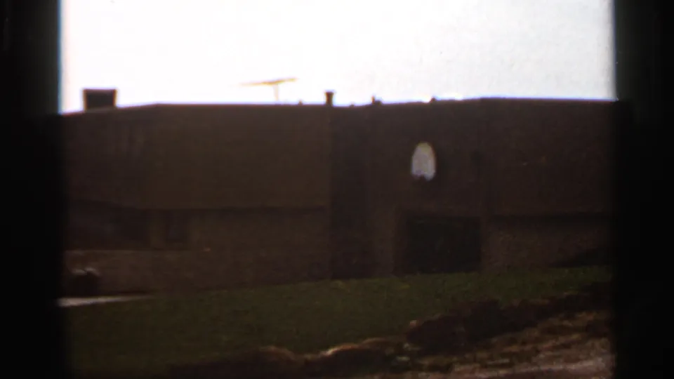 1966 8mm view in Saint Louis showing a dark building facade, roofline, small arched window, and grassy hill under a bright sky.