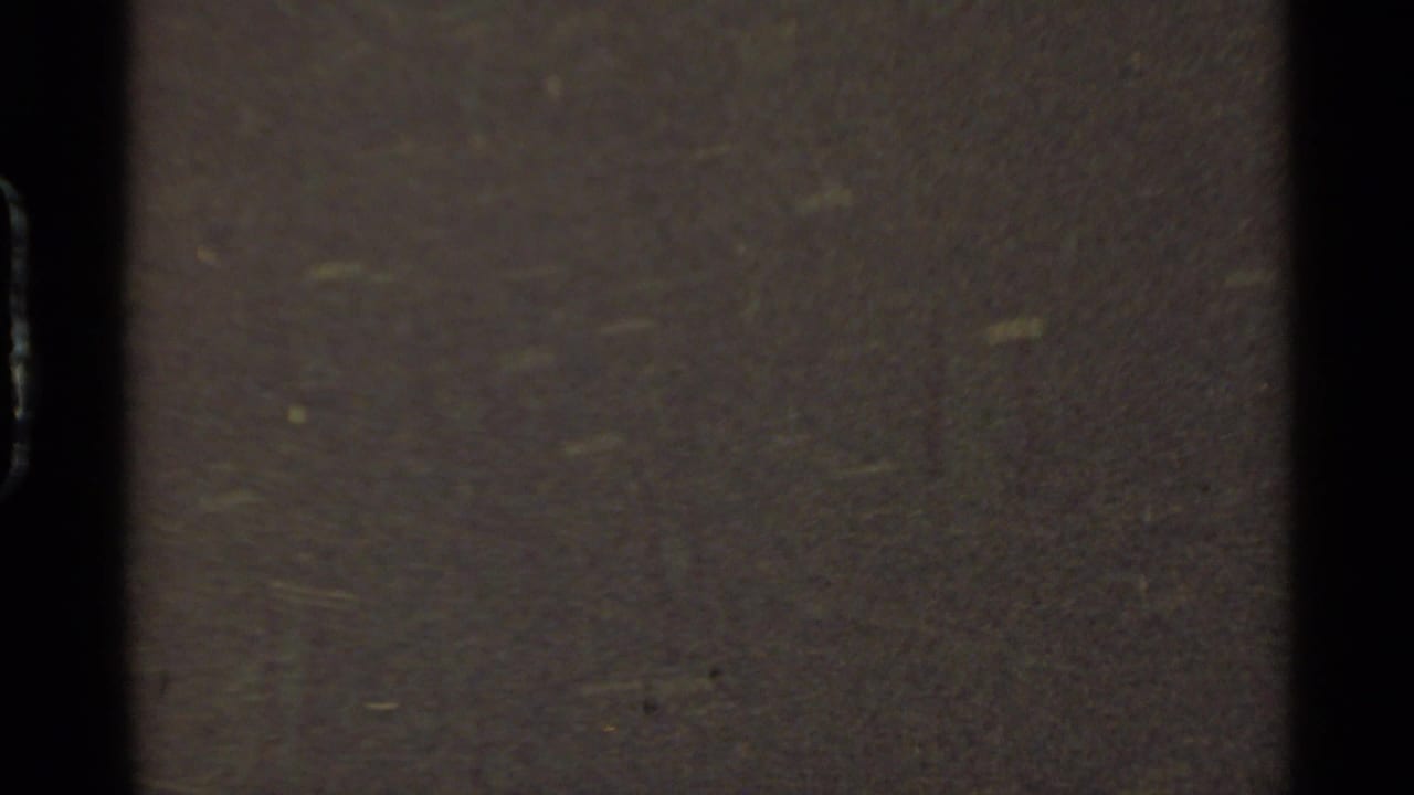 Final 8mm frame becomes an abstract close view of film surface texture, grain, dark exposure, and faint greenish specks.