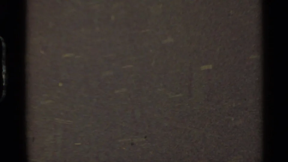 Final 8mm frame becomes an abstract close view of film surface texture, grain, dark exposure, and faint greenish specks.