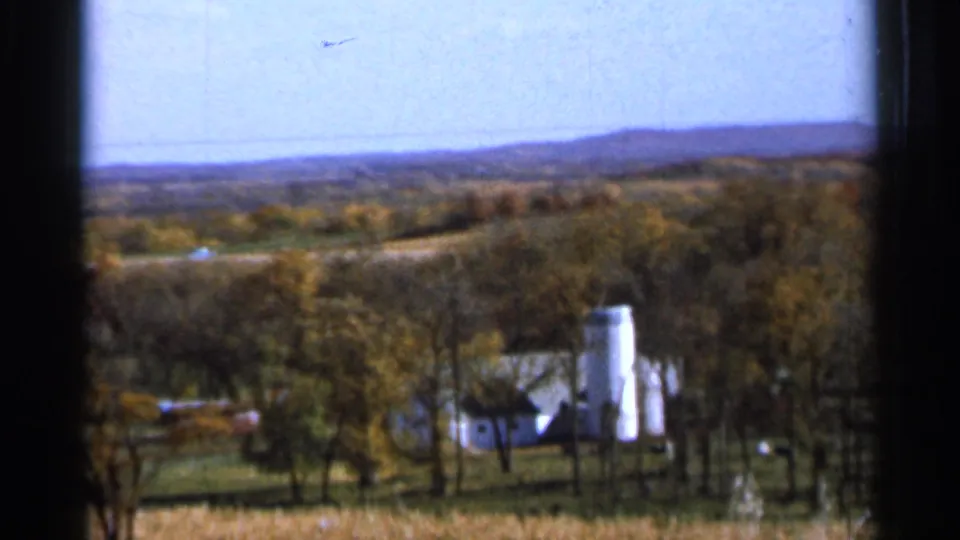 Soft 8mm frame of a Sauk Centre farmstead with white silo, surrounding trees, open land, and low distant hills.