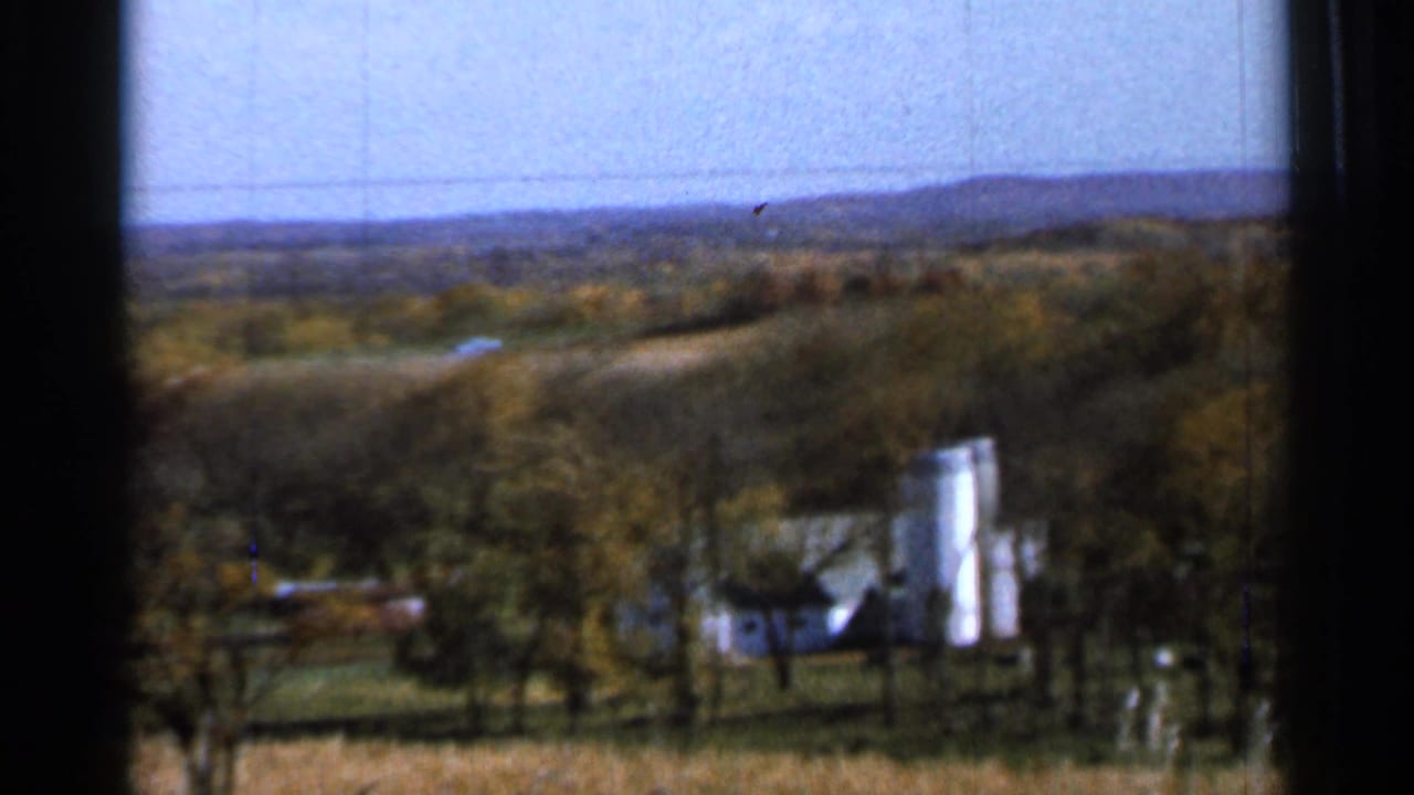 Distant farmhouse and silo seen across autumn fields through a vehicle window.