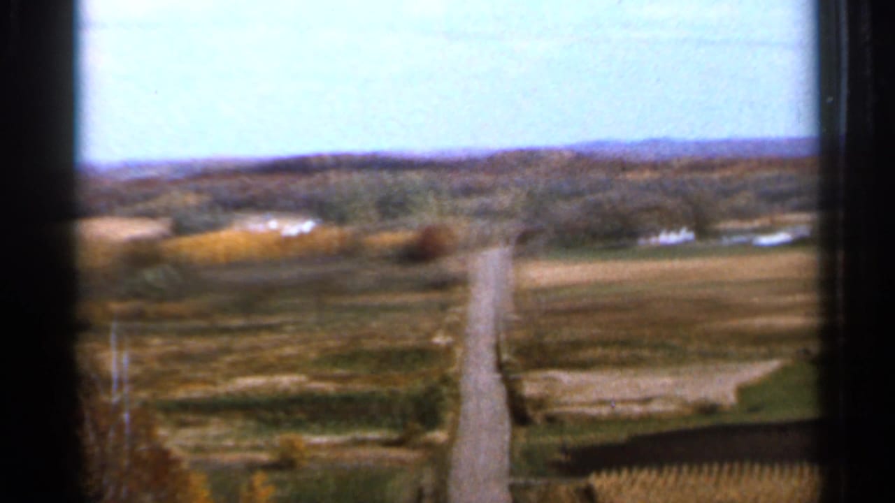 1966 Sauk Centre, Minnesota 8mm frame showing a long rural road, open fields, distant tree line, and small farm structures under bright sky.