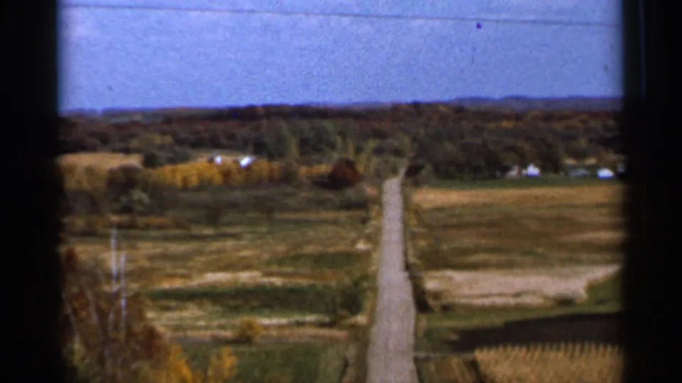1966 Sauk Centre, Minnesota view of a straight country road crossing green and golden fields toward distant houses and wooded horizon.