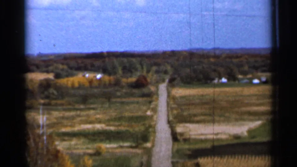 1966 Sauk Centre, Minnesota 8mm frame with open fields, a pale rural road, distant farm buildings, and autumn-toned trees at the horizon.