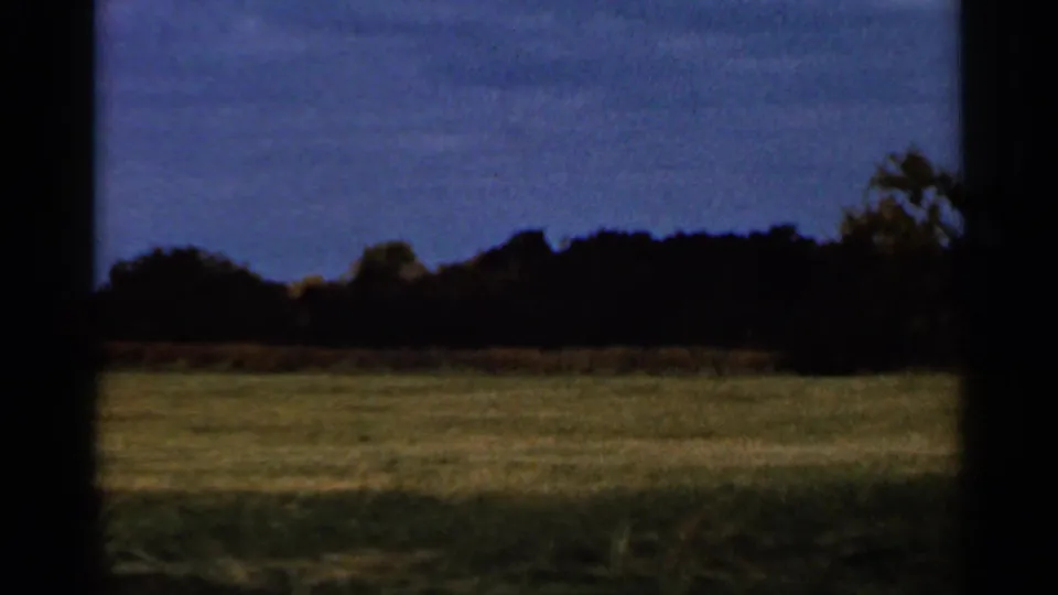 1966 Sauk Centre home-movie frame showing a grassy rural field and distant autumn tree line seen through dark window edges.