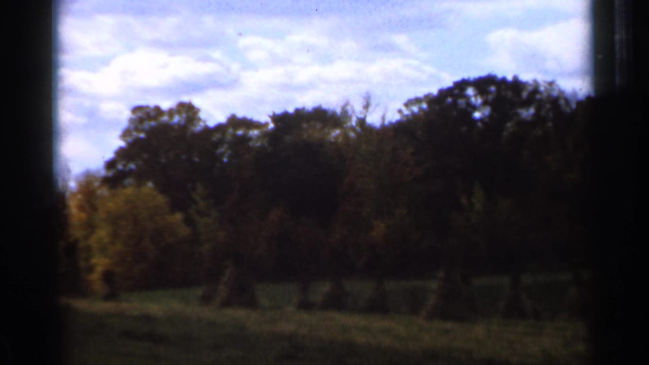 1966 Sauk Centre home-movie frame showing a quiet field with hay bales beneath a cloudy sky and autumn trees beyond.