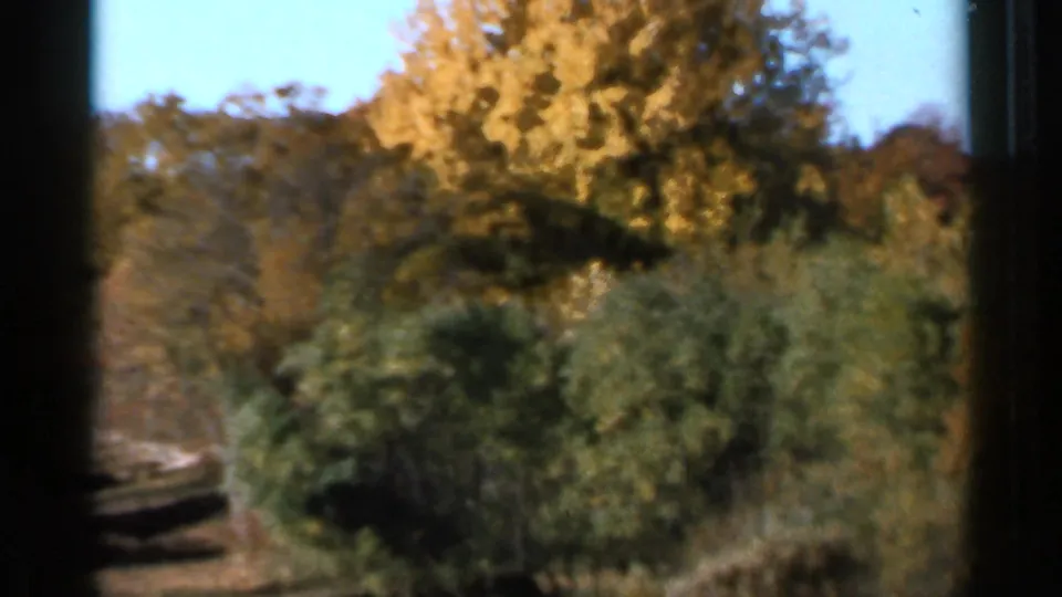 1966 Sauk Centre 8mm frame with yellow autumn trees, dense shrubs, blue sky, and a narrow path or road edge at left.