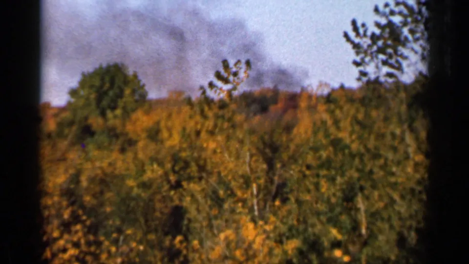 8mm home-movie view of a wooded autumn slope with dark smoke rising near the horizon in Sauk Centre, Minnesota.