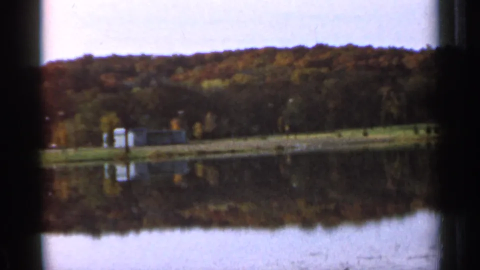 1966 Sauk Centre, Minnesota 8mm view of a still lake reflecting a wooded autumn hillside and small shoreline structures.