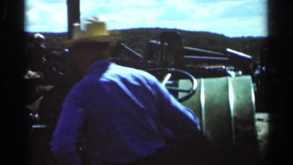 The 1966 rural Ohio home movie shows the tractor operator leaning over the controls of a green vintage machine.