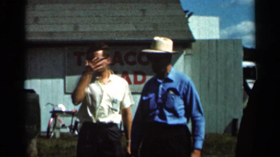 Two adult men near the steam engine area in Wauseon, Ohio, one gesturing while a Texaco sign, bicycle, and building appear behind them.