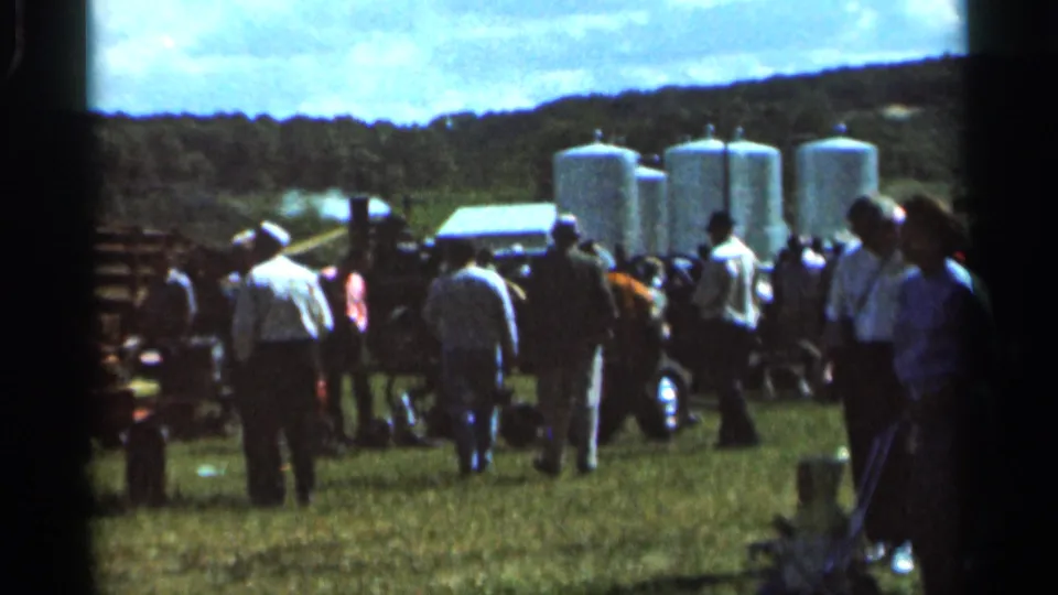 Spectators in 1960s dress move through a Wauseon field near machinery, white storage tanks, fencing, and parked vehicles.