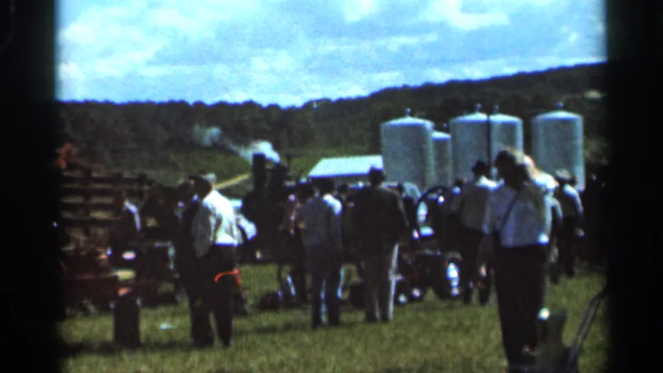 1966 Wauseon home-movie frame with people gathered near machinery, white tanks, fencing, and open countryside under a bright sky.