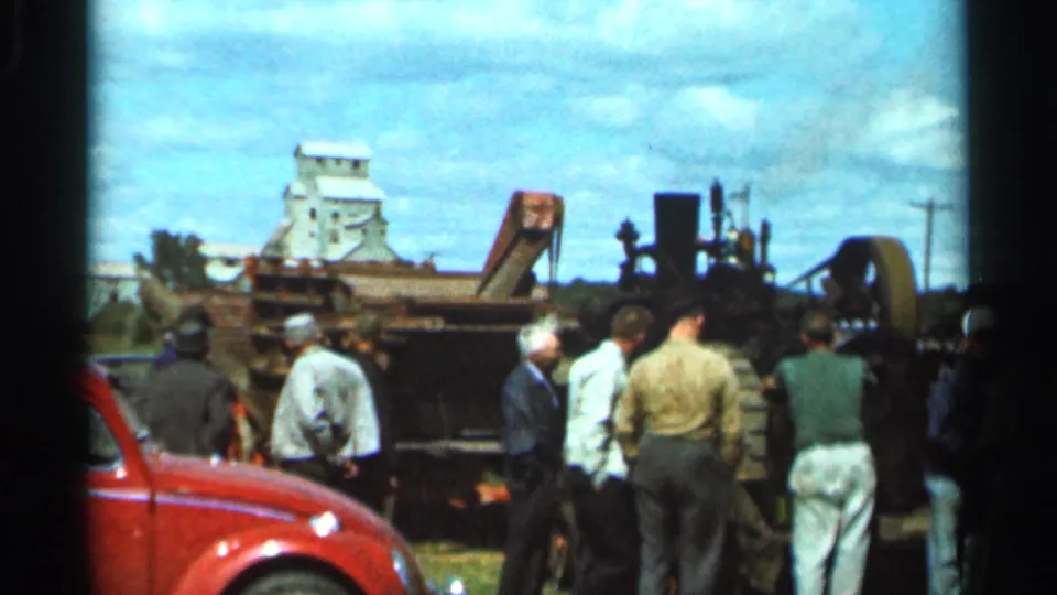 Wider 1966 Wauseon view with people watching rail equipment near a red car and grain elevator under a cloudy sky.
