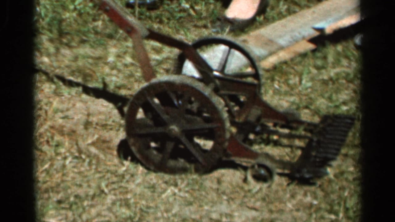 Close view of a metal push mower on dry grass with spoked wheels and a partial foot nearby.