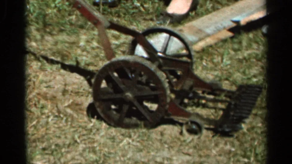 1966 Wauseon, Ohio home-movie frame showing a simple mechanical push mower resting on dry grass, with spoked wheels and a partial foot visible.