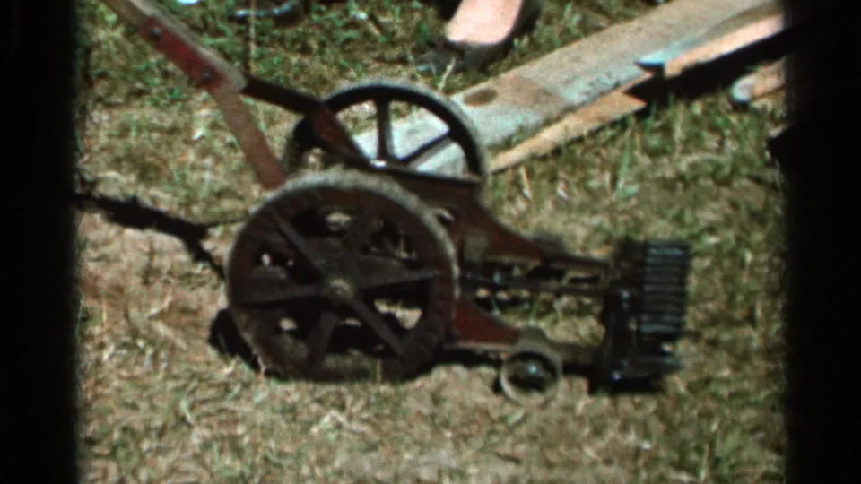 Close 1966 home-movie view of a mechanical lawn mower in Wauseon, Ohio, showing its wheels, handle, and blade assembly on dry grass.