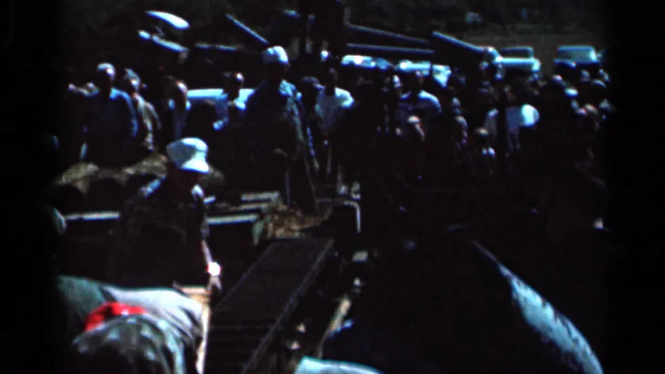 Dark 8mm frame from the same 1966 Wauseon, Ohio clip showing people gathered outdoors near vehicles, with faces mostly indistinct.