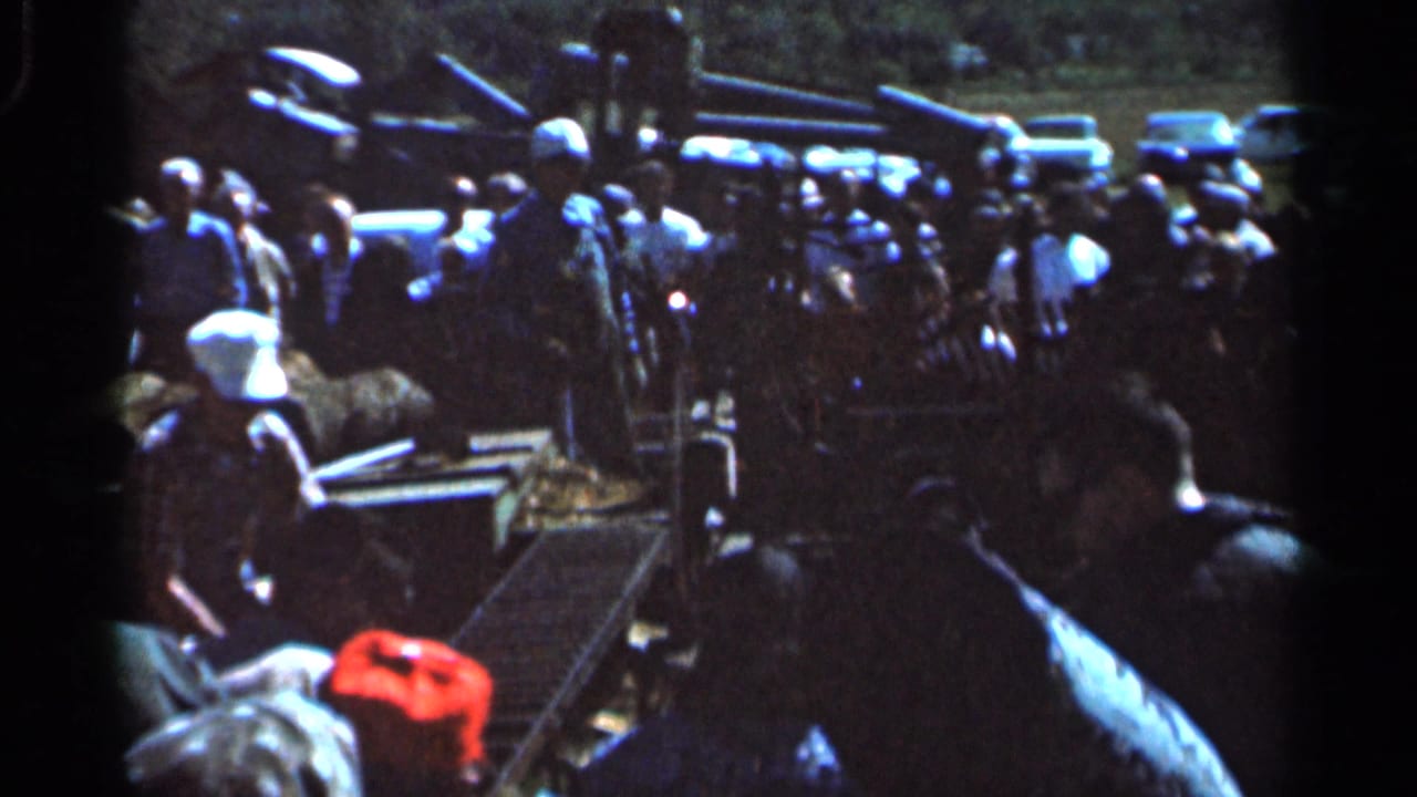 1966 Wauseon, Ohio home-movie frame showing adults gathered around field equipment with cars and trees in the background.