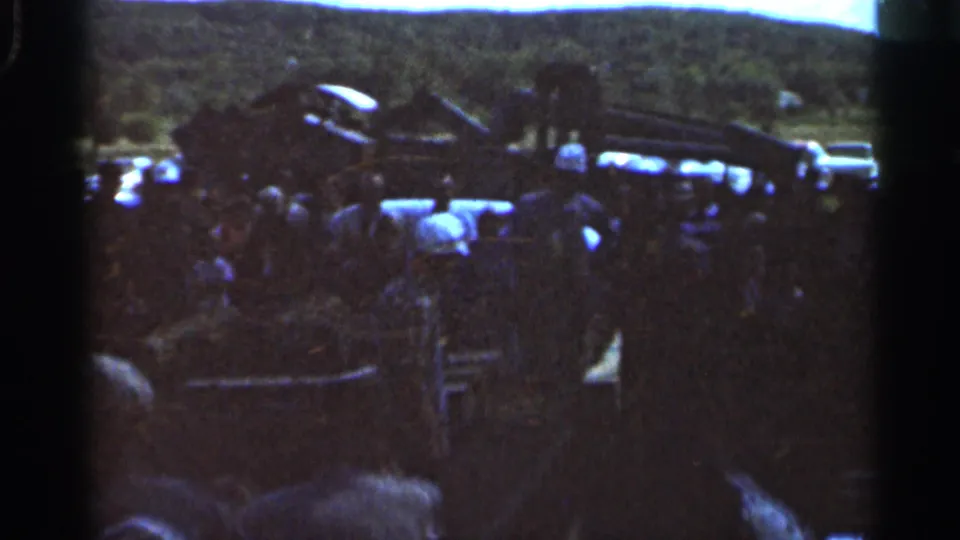 Home-movie frame from the 1966 Wauseon scene showing adults near a ladder or conveyor structure, machinery, and vehicles.