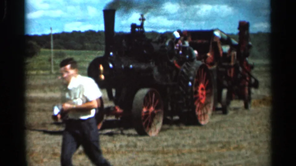 In 1966 Wauseon, Ohio footage, a young man in a white shirt passes a black steam tractor with red wheels in a rural field.