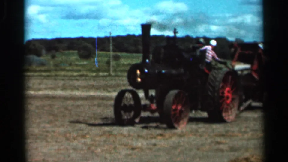 A man in a plaid shirt and white hat stands near the red-wheeled steam tractor during the 1966 Wauseon, Ohio footage.