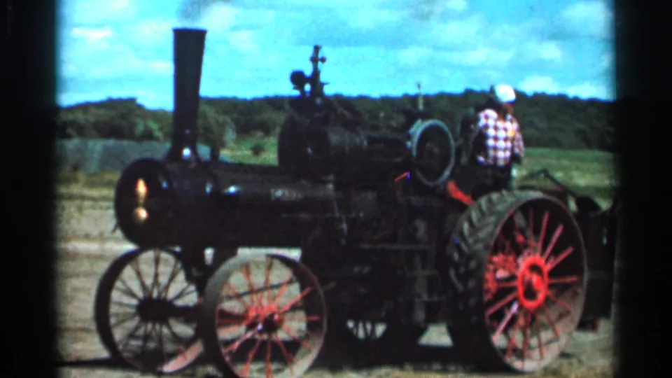 Black steam tractor in Wauseon, Ohio, with a plaid-shirted operator near the rear wheel during a field demonstration.