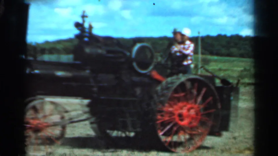 Closer 8mm view of the steam tractor's side, red rear wheel, and operator area in a Wauseon field.