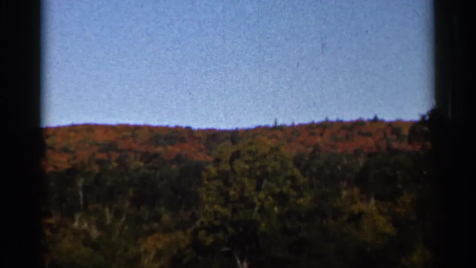 1966 Wauseon, Ohio 8mm frame showing a distant wooded hillside with red, orange, and green fall foliage below a blue sky.