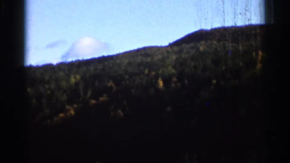 1966 8mm frame of a dark tree-covered slope beneath pale blue sky, with a small cloud and visible film wear.