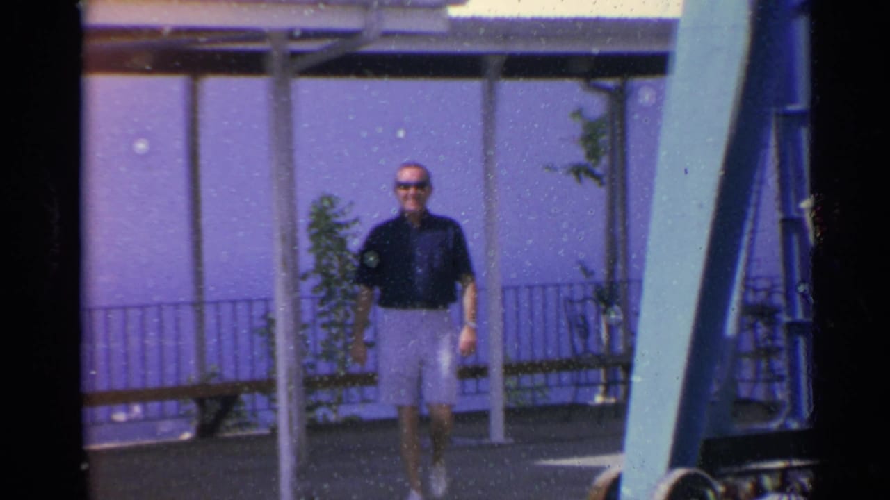 1967 US Virgin Islands home-movie frame showing a man in sunglasses walking near a seaside amusement ride and railing.