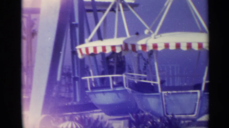 1967 US Virgin Islands home-movie frame focused on blue gondola seats and striped canopies near a seaside boardwalk railing.