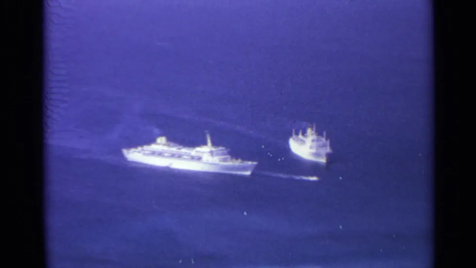 Distant 1967 home-movie view of a larger white ship and a smaller vessel crossing blue sea under soft exposure.