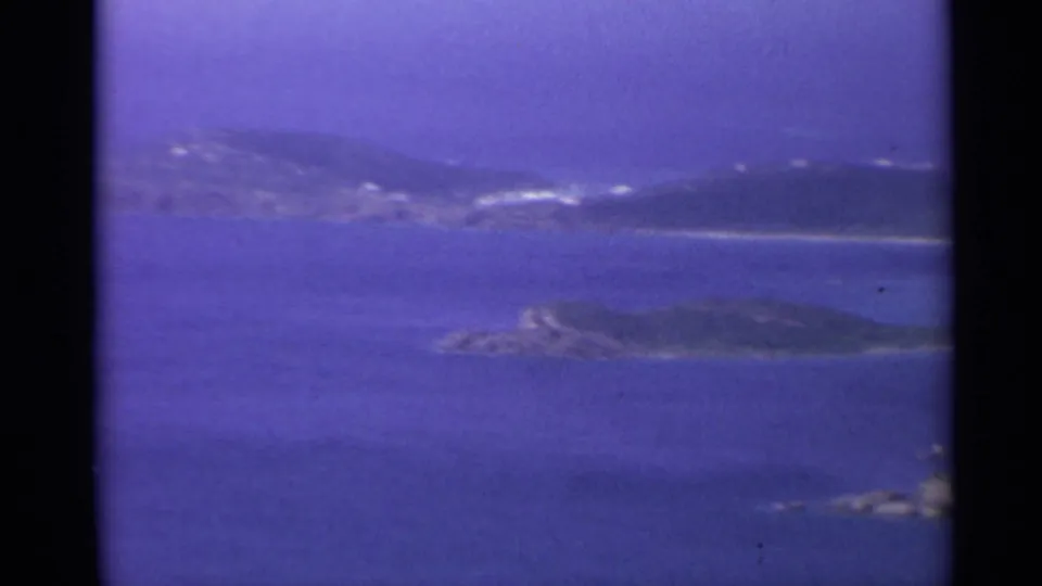 1967 U.S. Virgin Islands shoreline view with green hills, blue channels, rocky foreground islands, and small white buildings in the distance.