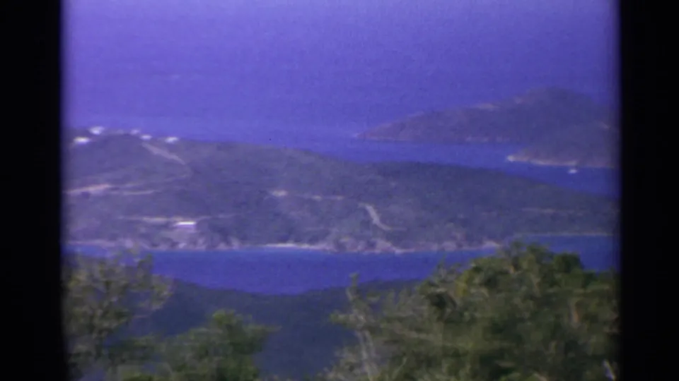 1967 8mm travel view over a U.S. Virgin Islands bay with a green ridge, distant island, and foreground foliage.