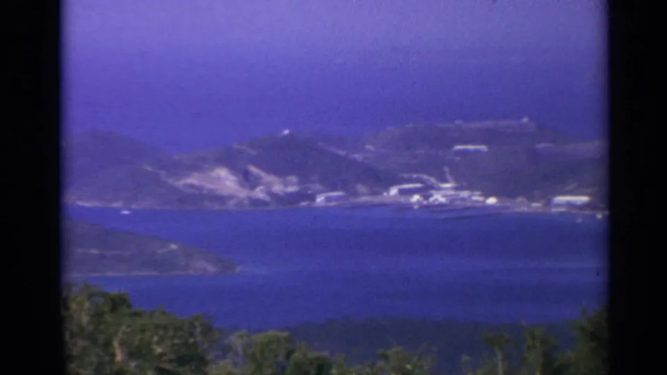 Soft 1967 8mm view across the water toward a US Virgin Islands shoreline with low hills, pale buildings, and foreground vegetation.
