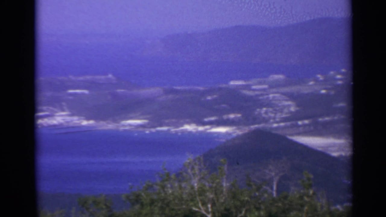 Elevated view of blue water, a coastal settlement, green hills, and distant mountains.