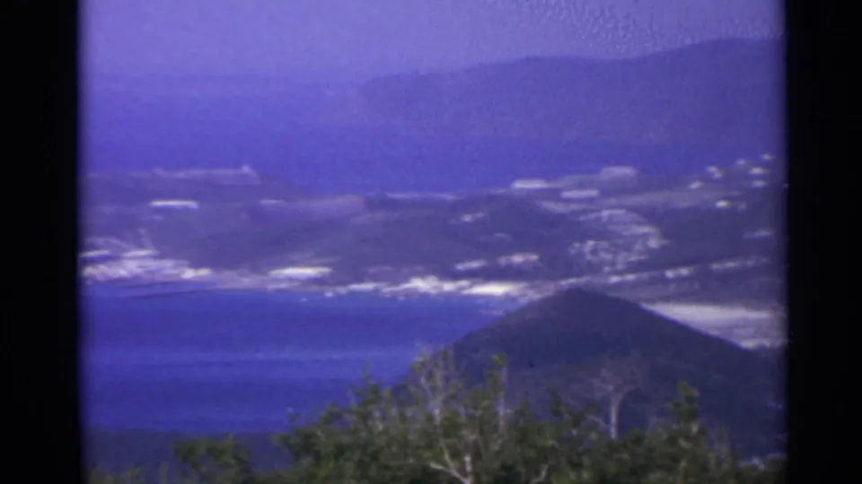 1967 8mm view likely in the US Virgin Islands, showing a coastal town below mountains with blue water and foreground foliage.