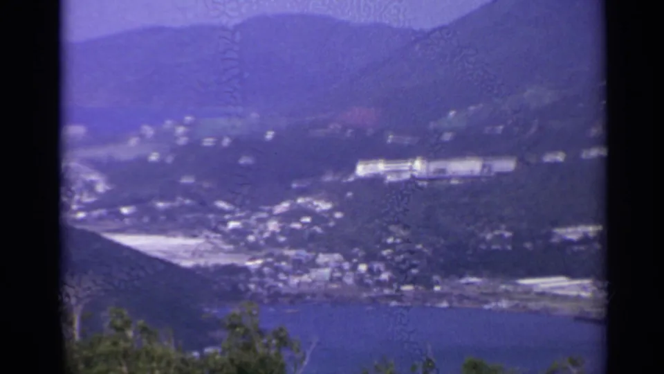 Soft 8mm frame likely from the US Virgin Islands, showing clustered shoreline buildings below hazy mountain slopes.