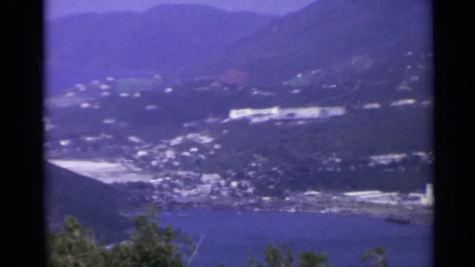 1967 island landscape frame with harbor water, hillside settlement, pale buildings, and mountains under a purple-blue film cast.