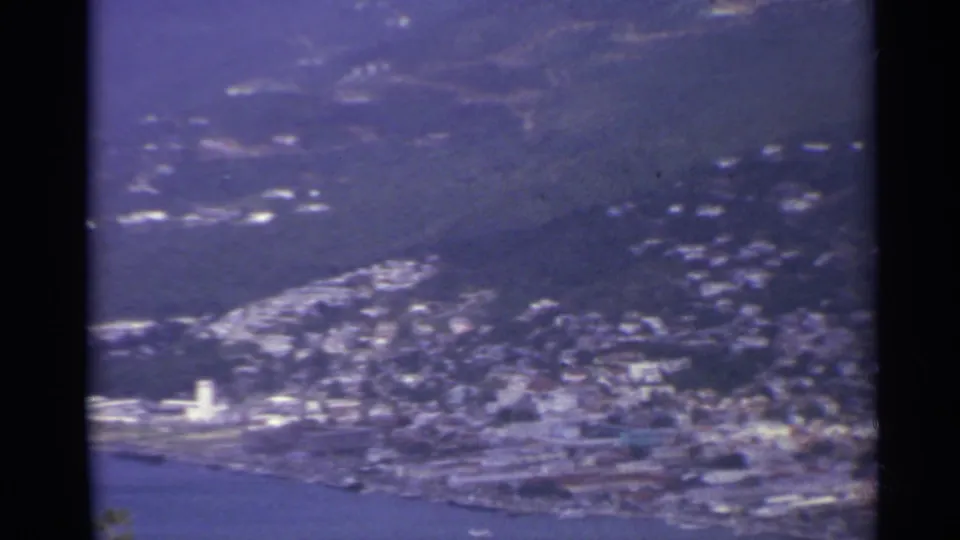 1967 US Virgin Islands shoreline view with a tall white tower, dense hillside housing, waterfront roads, and blue bay water.