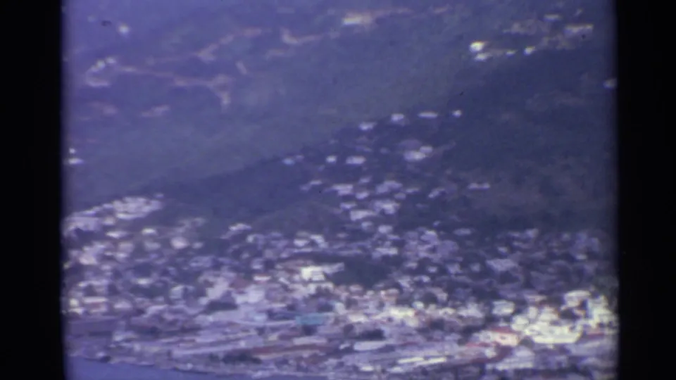 Soft 8mm frame of a US Virgin Islands hillside town in 1967, with clustered pale buildings above a waterfront and bay.