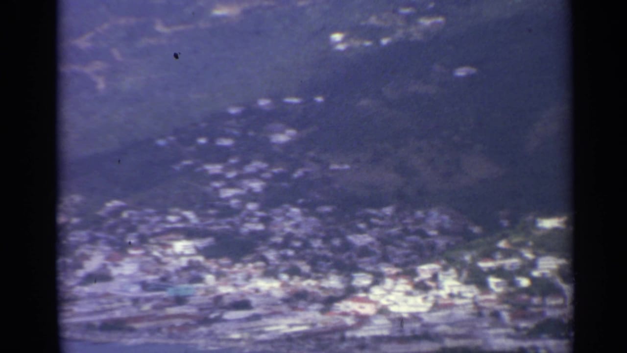 Soft aerial view of a coastal town below green hills and dark frame edges.